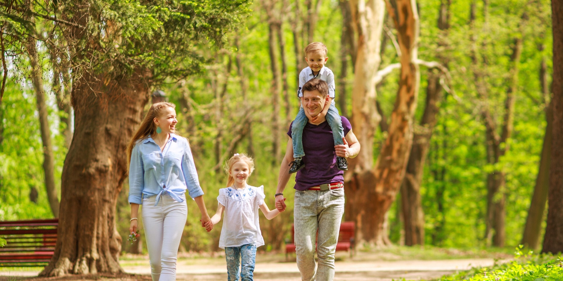Een vrolijk gezin maakt een wandeling door een zonnig bos; de ouders houden de hand van hun dochtertje vast, terwijl hun zoontje op de schouders van de vader zit.